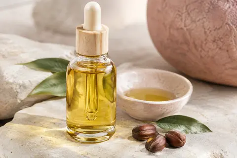 Flacon en verre d’huile de jojoba posé sur une pierre claire à côté d’une coupelle en céramique.