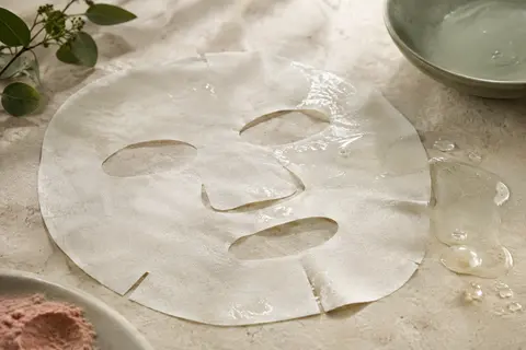 Masque visage en tissu et texture de sérum présentés sur une pierre claire, dans une lumière douce.