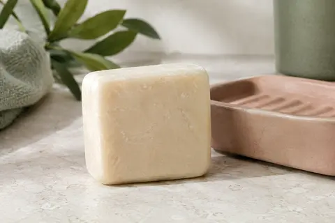 Savon solide crème au lait de riz posé sur une pierre claire près d’un porte-savon en céramique.