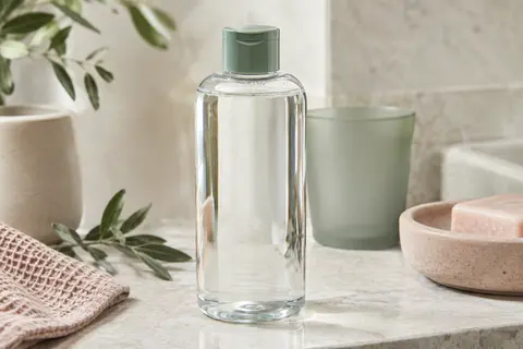 Flacon d’eau micellaire pour peaux sensibles posé sur une pierre claire, près d’un verre dépoli.
